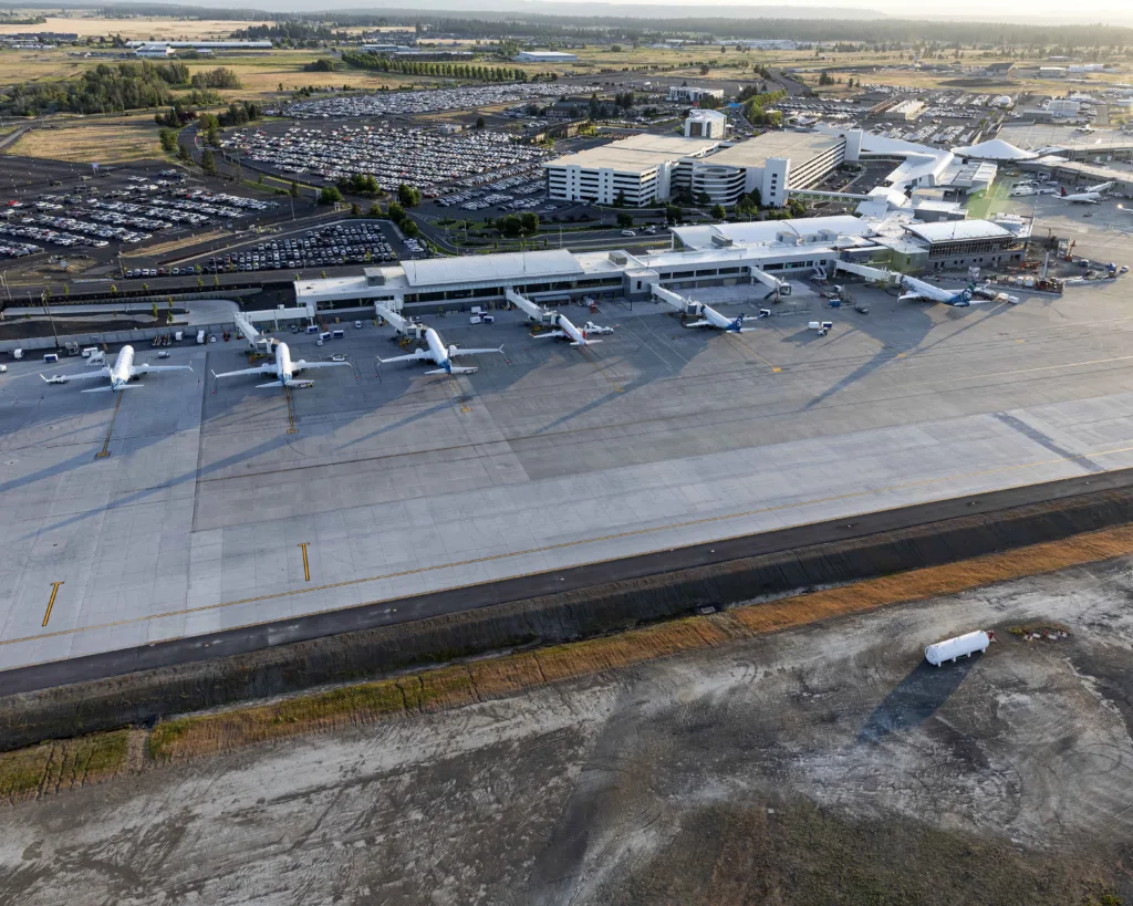 An aerial photograph of Spokane International Airport showing multiple commercial aircraft parked at gates along the terminal, a large tarmac apron, expansive parking lots, the terminal and parking structure, and the surrounding landscape under warm evening light.