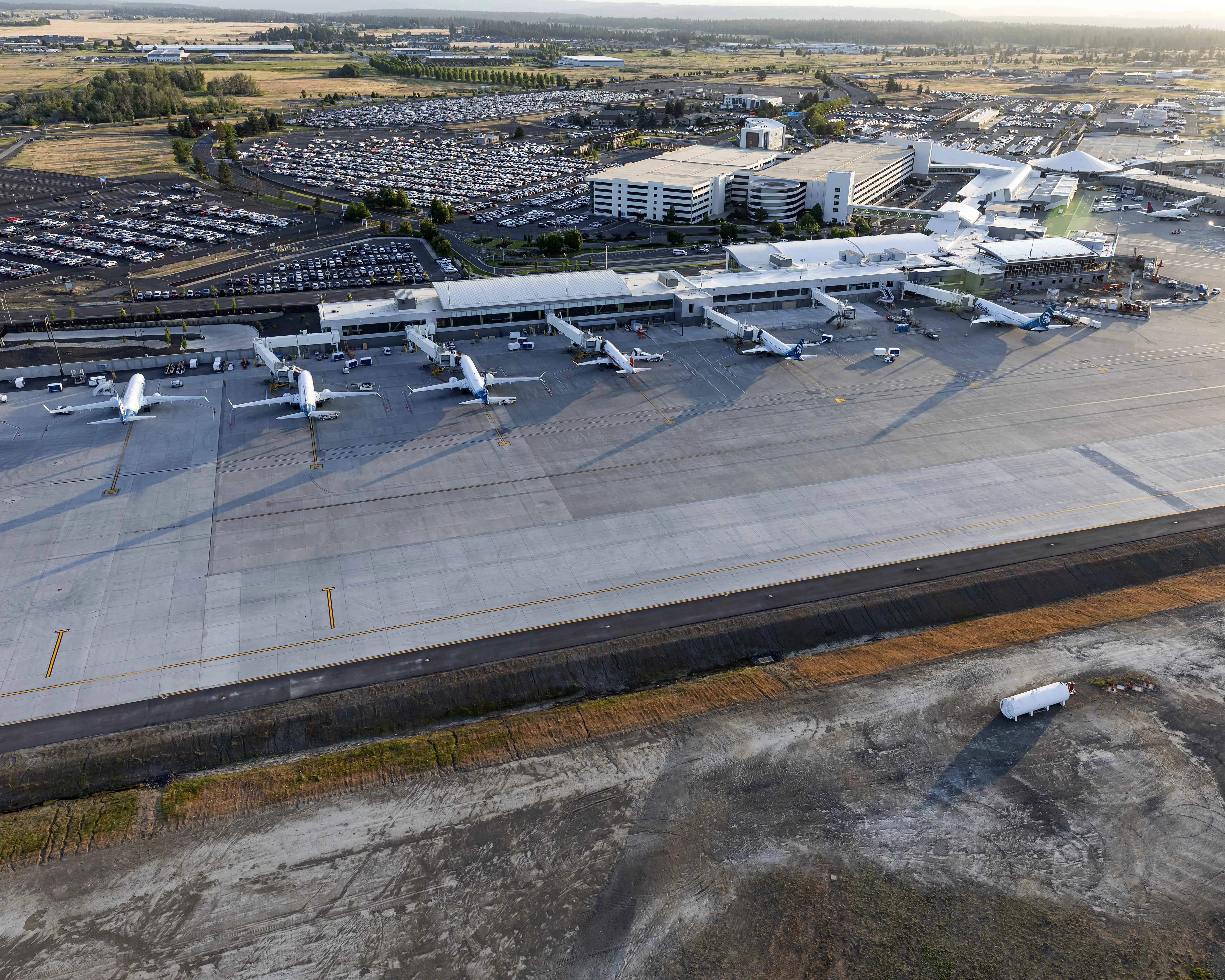 An aerial photograph of Spokane International Airport showing multiple commercial aircraft parked at gates along the terminal, a large tarmac apron, expansive parking lots, the terminal and parking structure, and the surrounding landscape under warm evening light.