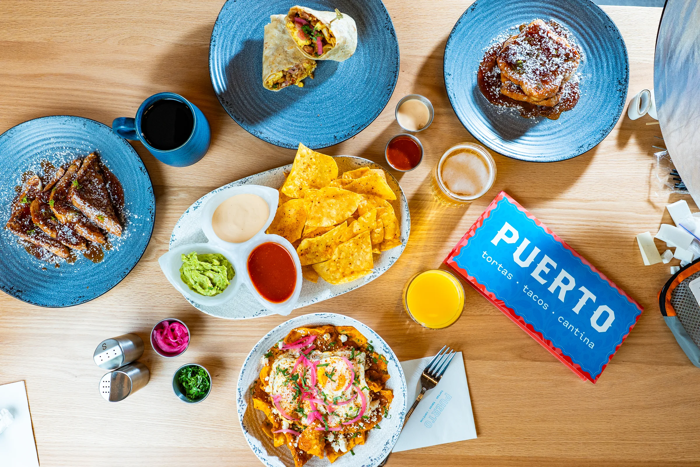 An overhead flat-lay of a spread of food and drinks from Puerto Tortas Tacos Cantina, including chilaquiles topped with a fried egg and pickled onions, chips with guacamole and salsa, a breakfast burrito, French toast, pancakes dusted with powdered sugar, orange juice, coffee, and a beer, arranged on blue plates on a wooden table alongside the Puerto branded menu.