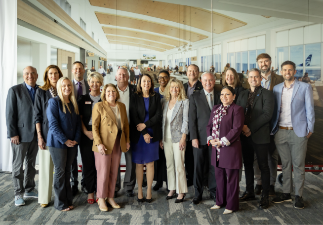 A group of airport officials, project stakeholders, and community leaders pose together inside the newly completed Concourse C at Spokane International Airport, with the modern terminal interior and an Alaska Airlines aircraft visible in the background.