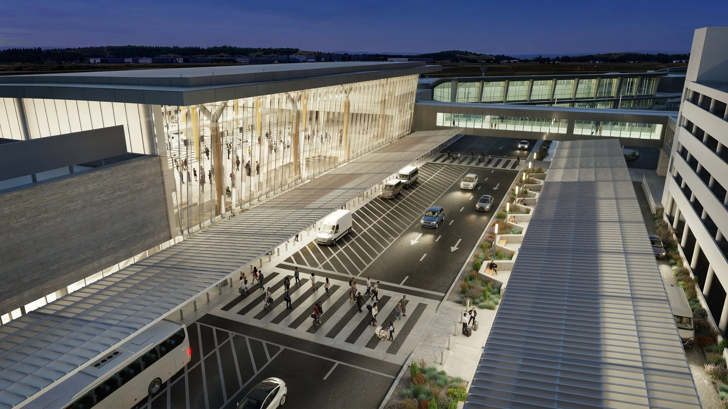 An aerial architectural rendering of a modern airport central hall at dusk, showing a glowing glass-facade terminal building, a multi-lane passenger drop-off and pick-up roadway with vehicles and pedestrians, a covered walkway, and a parking structure to the right.