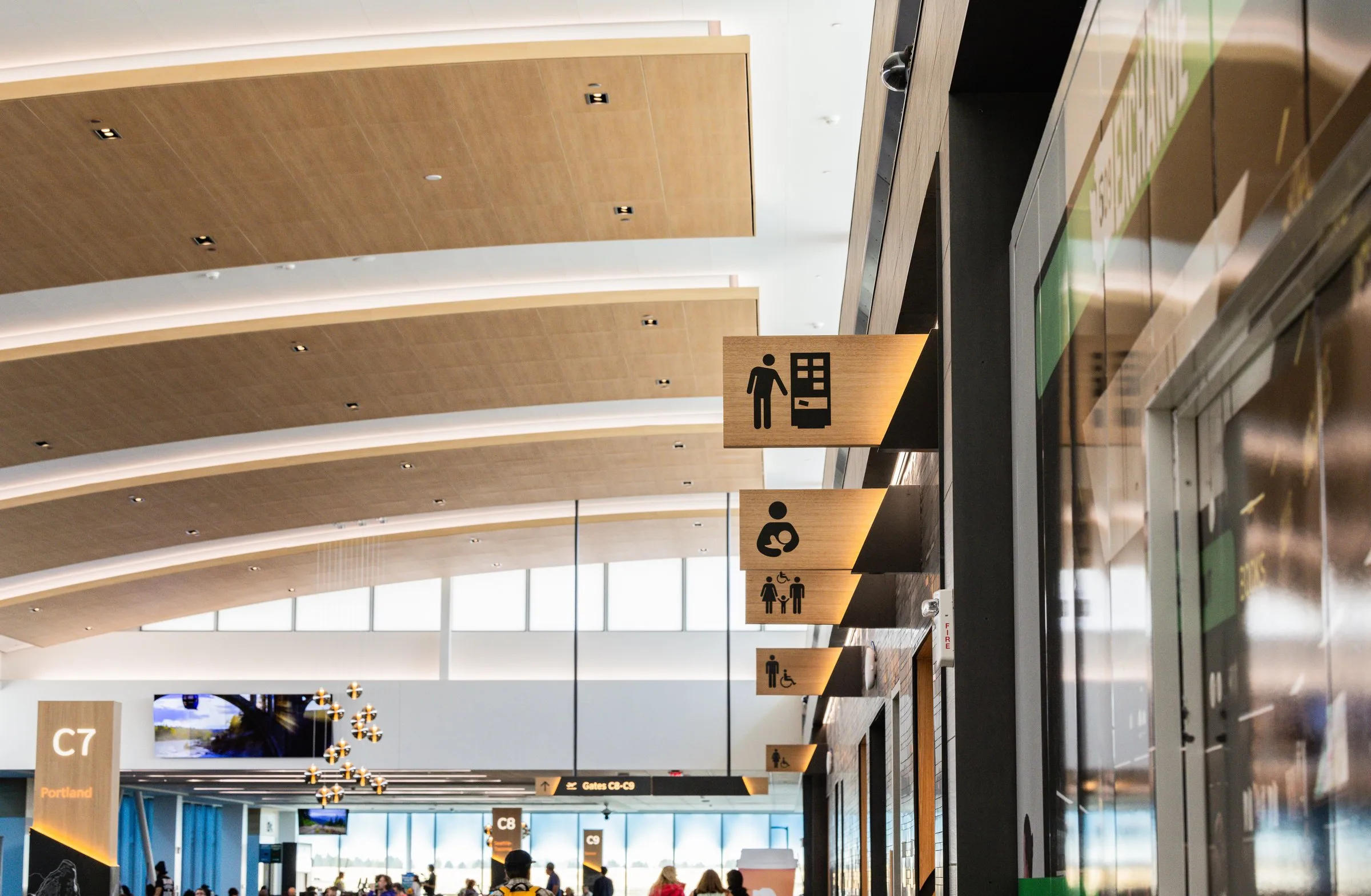 A close-up of warm wood-toned wayfinding signage in Concourse C at Spokane International Airport, displaying icons for a vending machine, lactation room, family restroom, and accessible restroom, with gate signage for C7 Portland, C8, and C9 and a busy concourse visible in the background.