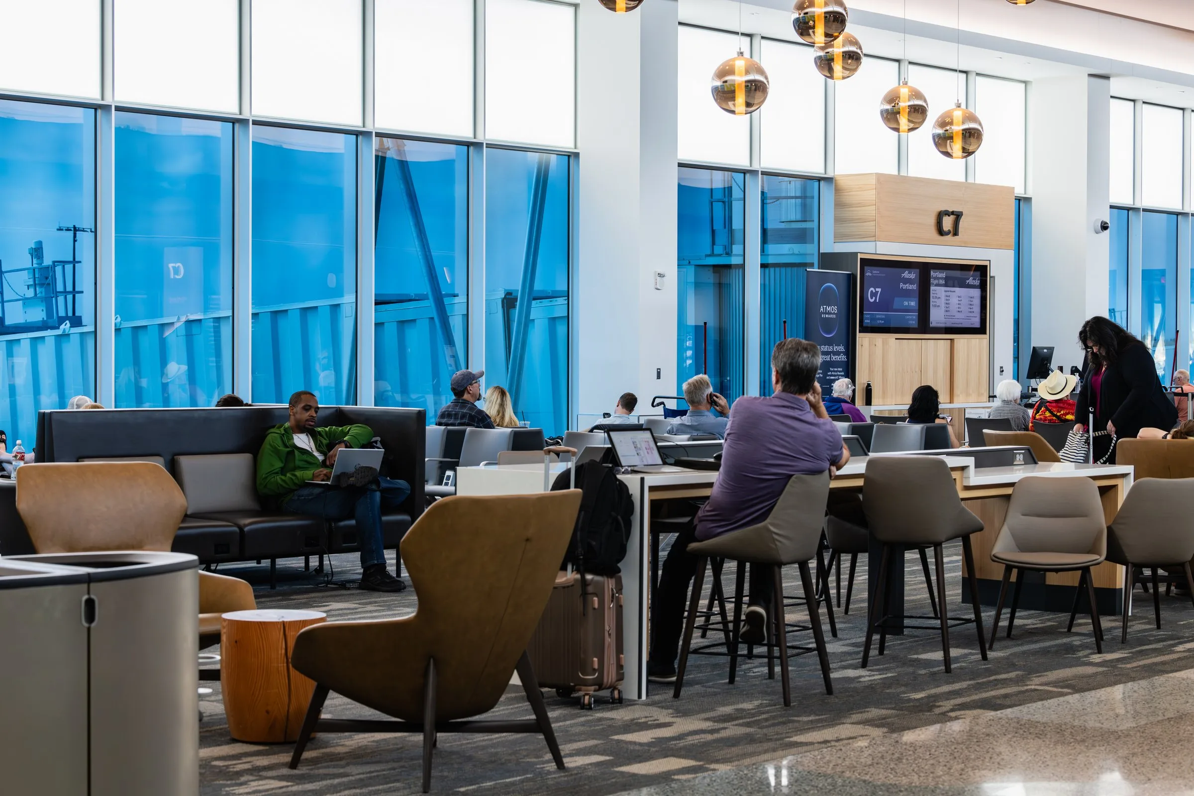 Travelers work on laptops and relax in a modern gate seating area at Gate C7 in Concourse C at Spokane International Airport, featuring mixed seating including lounge chairs, booth seating, and a work counter with bar-height chairs, globe pendant lighting, floor-to-ceiling windows, and a flight information display screen.