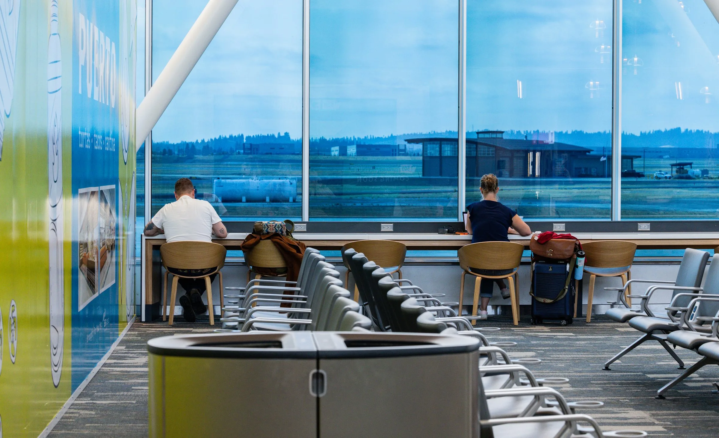 Two travelers seated at a window-side work counter in Concourse C at Spokane International Airport, with floor-to-ceiling windows overlooking the airfield and a colorful mural wall to the left.