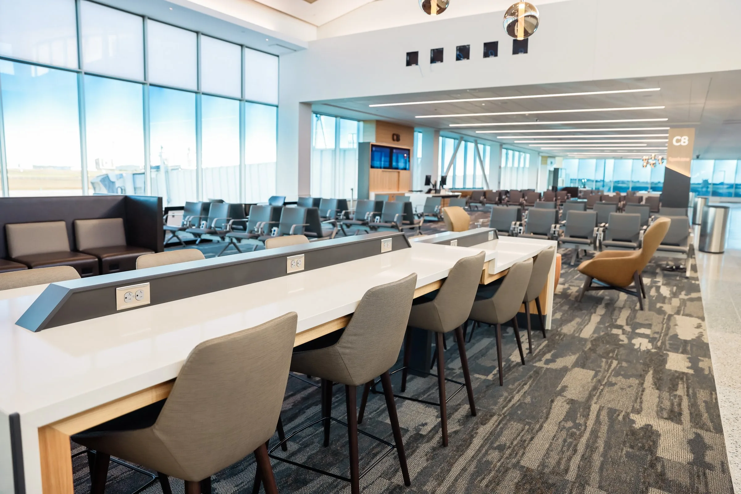 A bright, open gate area in Concourse C featuring a long white work counter with built-in power outlets, bar-height seating, rows of gate chairs, accent lounge chairs, and floor-to-ceiling windows overlooking the airfield.