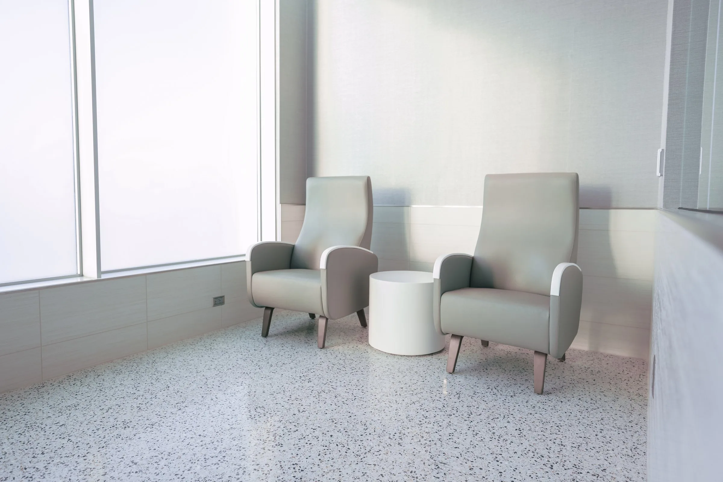 Two high-back upholstered armchairs in soft gray tones flank a small round white side table in a bright, minimalist airport lounge seating area with terrazzo flooring and frosted windows.