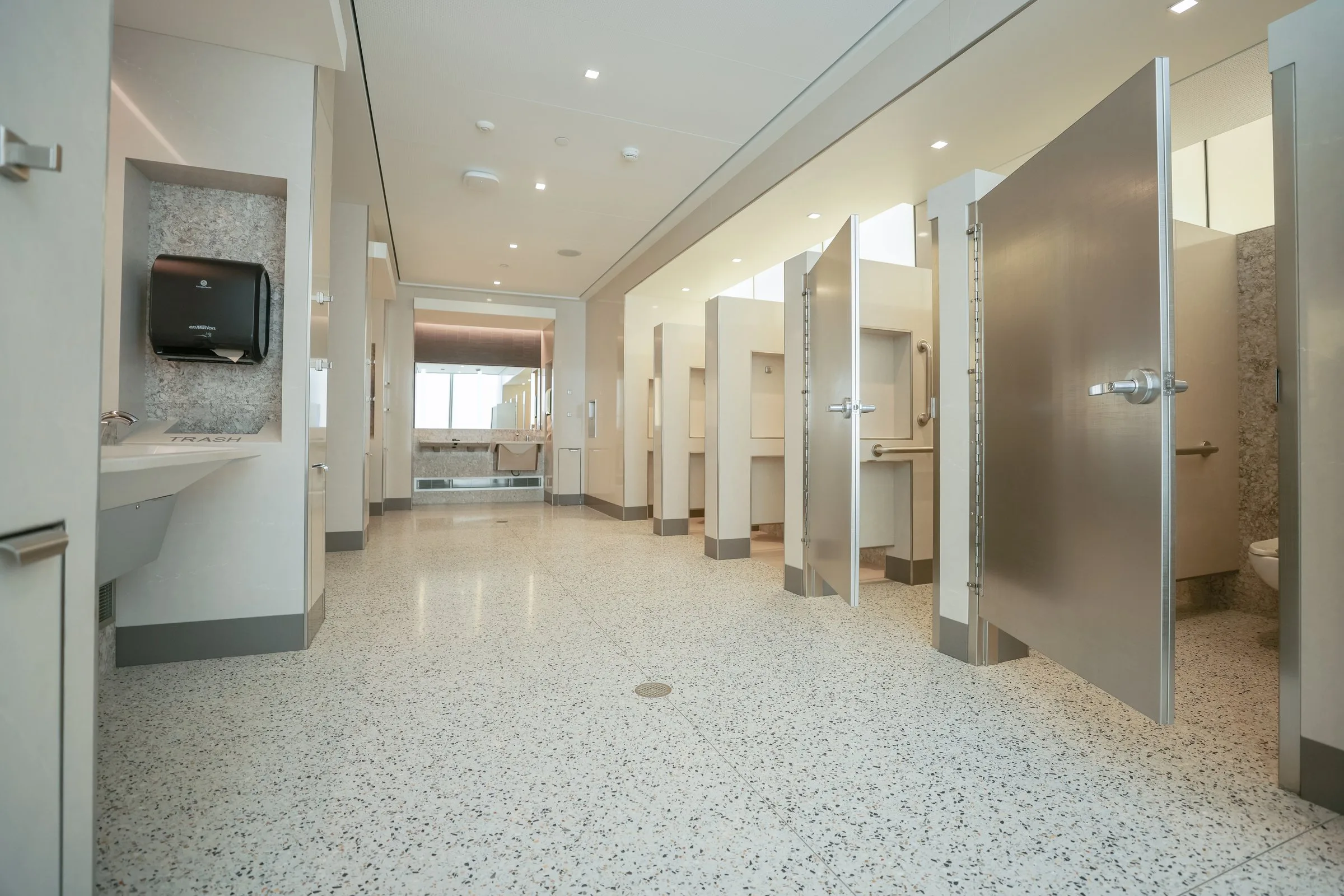A spacious, modern public restroom with terrazzo floors, stainless steel stall partitions, recessed lighting, and accessible stalls with grab bars.