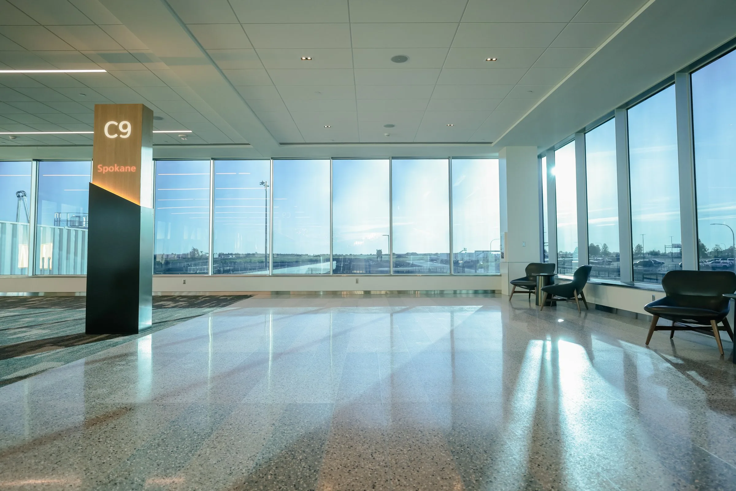 A bright, open gate area at Gate C9 with polished terrazzo floors, floor-to-ceiling windows overlooking the airfield, a wooden wayfinding pylon displaying "C9 – Spokane," and a small cluster of dark lounge chairs along the window wall.