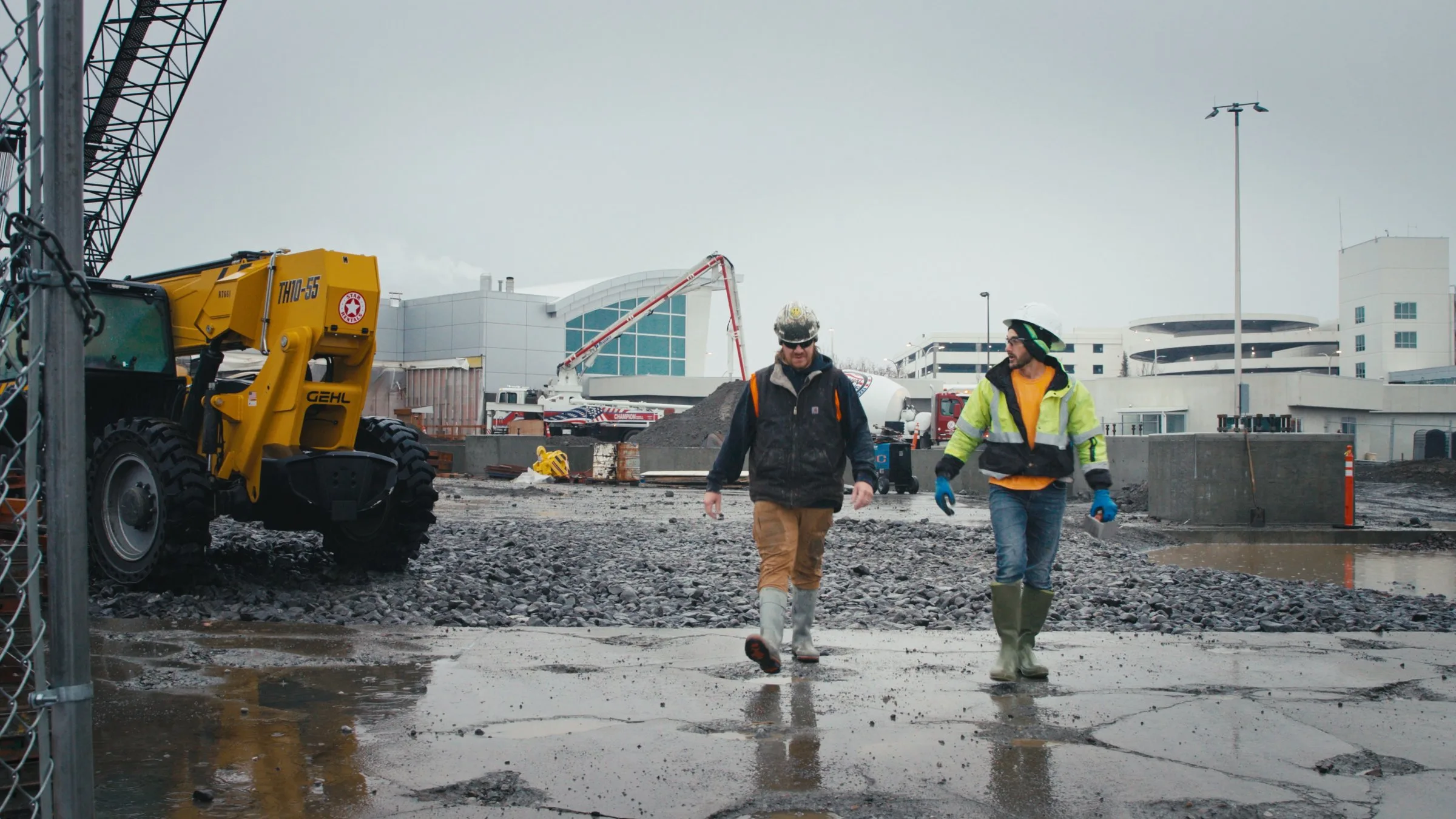 Two construction workers in hard hats and high-visibility safety gear walk across a muddy, gravel-covered construction site with heavy equipment and airport terminal buildings visible in the background.