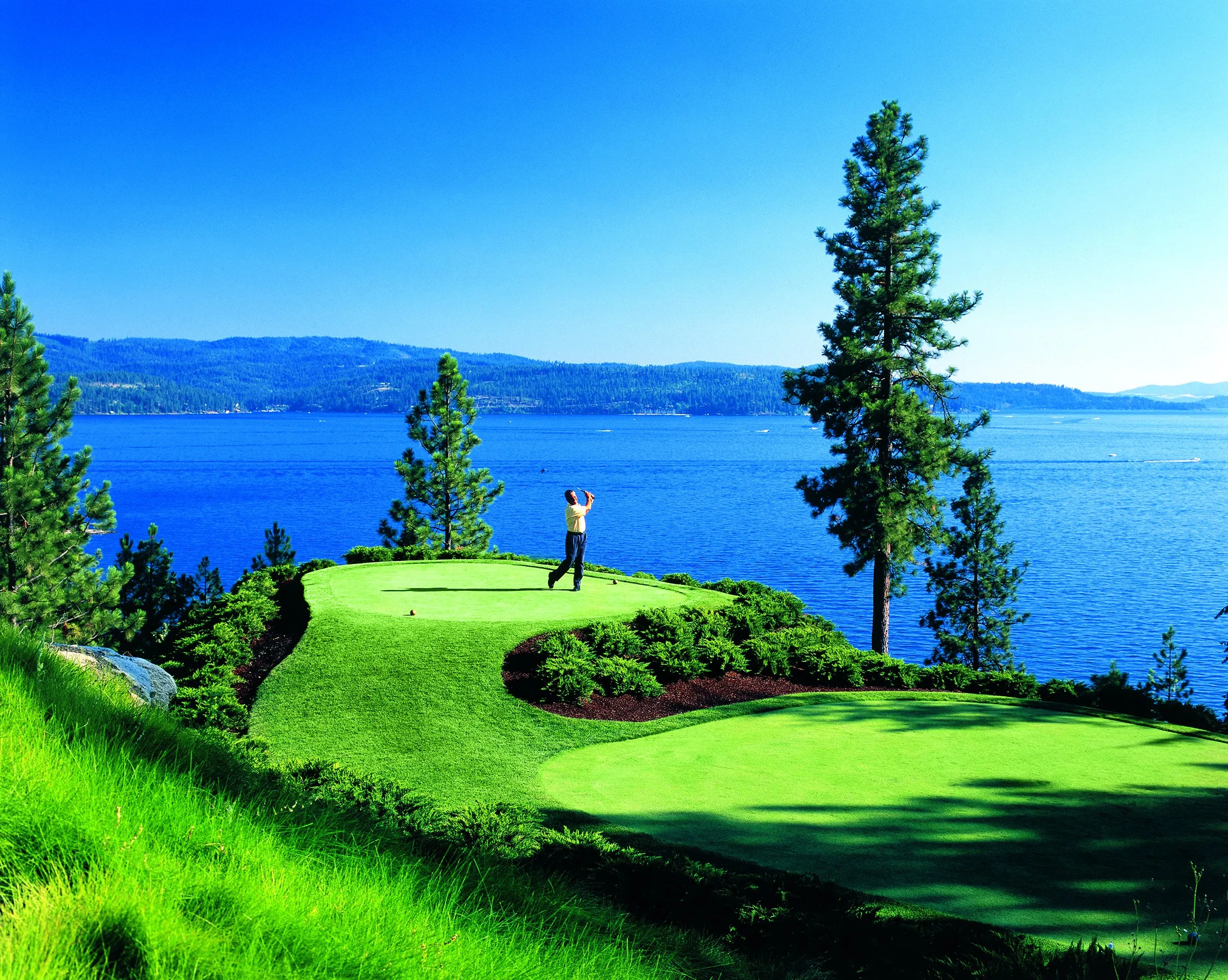 A golfer finishes a swing on a lush green golf course hole perched above the deep blue waters of Lake Coeur d'Alene, surrounded by tall pine trees and manicured landscaping under a clear blue sky.