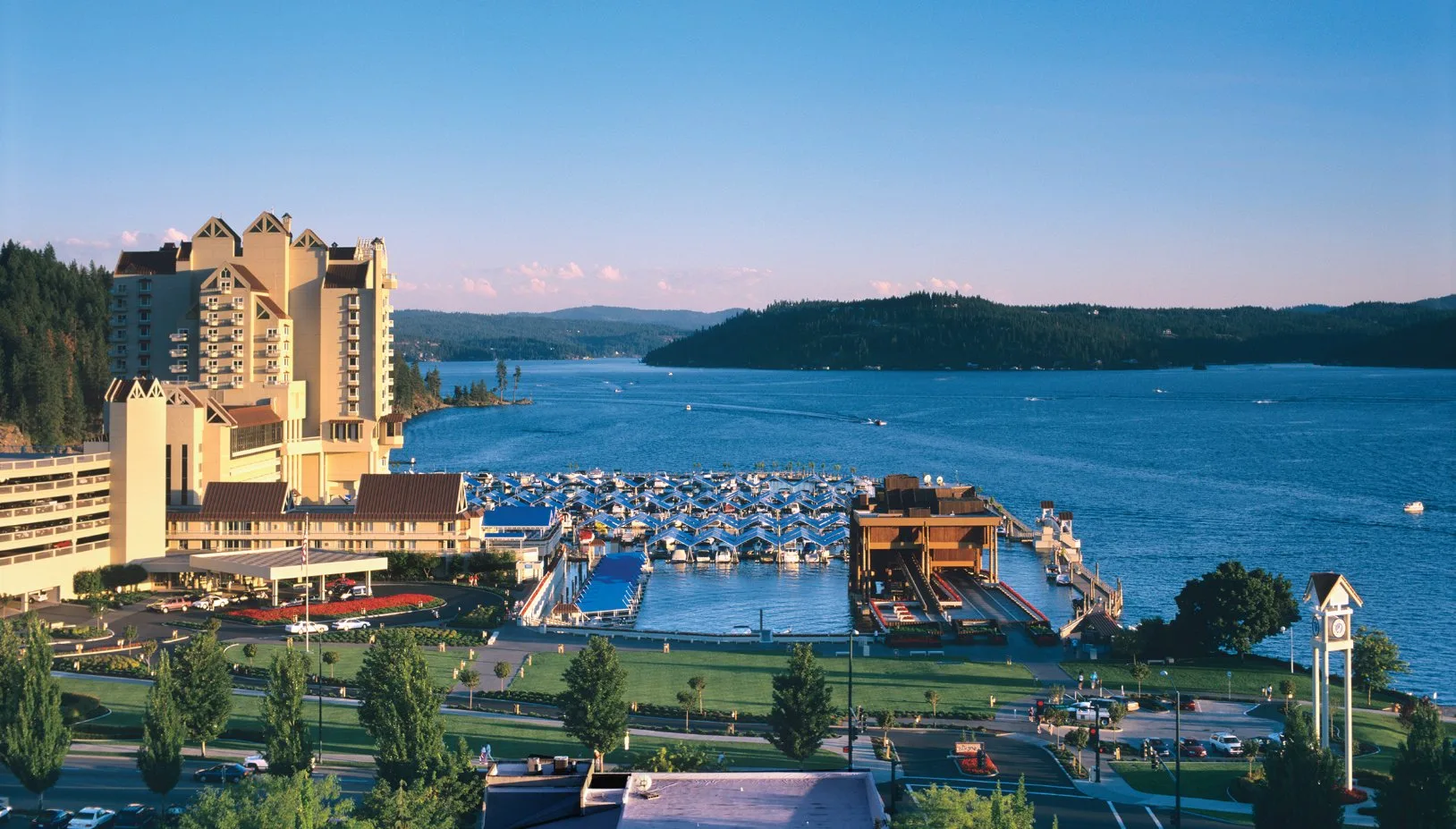 Aerial view of the Coeur d'Alene Resort and its marina on Lake Coeur d'Alene in northern Idaho, with forested hillsides, open water, and the resort's distinctive peaked roofline visible against a clear blue sky.