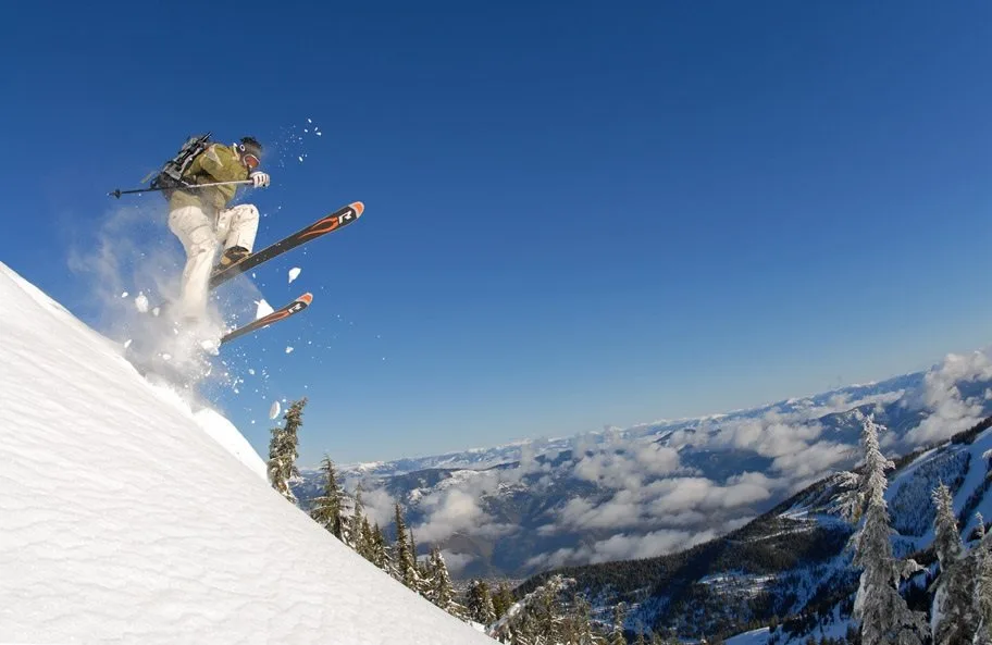 A skier launches off a steep snow-covered slope at Silver Mountain Resort, with sweeping mountain valley views, snow-dusted evergreen trees, and a clear blue sky in the background.
